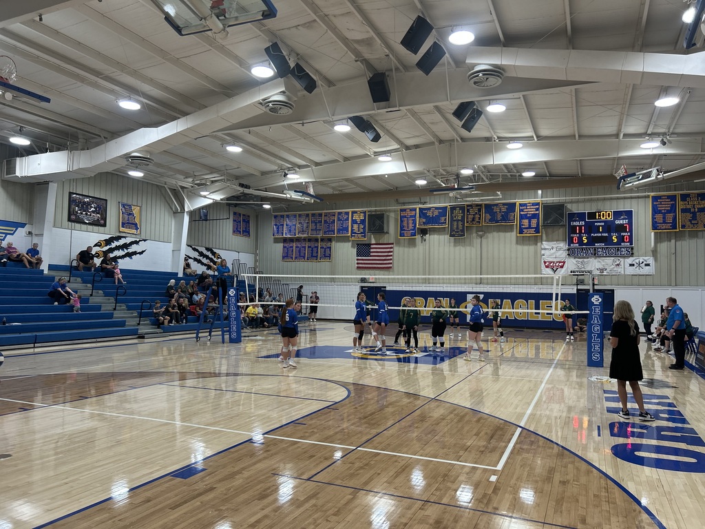 wo volleyball teams gather near the net on a polished school gym court as spectators watch from the bleachers.