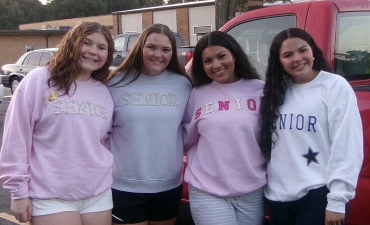 Four students in pastel “Senior” sweatshirts pose together in a parking lot beside a red vehicle.