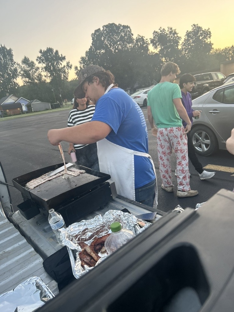 A student wearing an apron cooks strips of bacon on an outdoor griddle as classmates gather nearby.