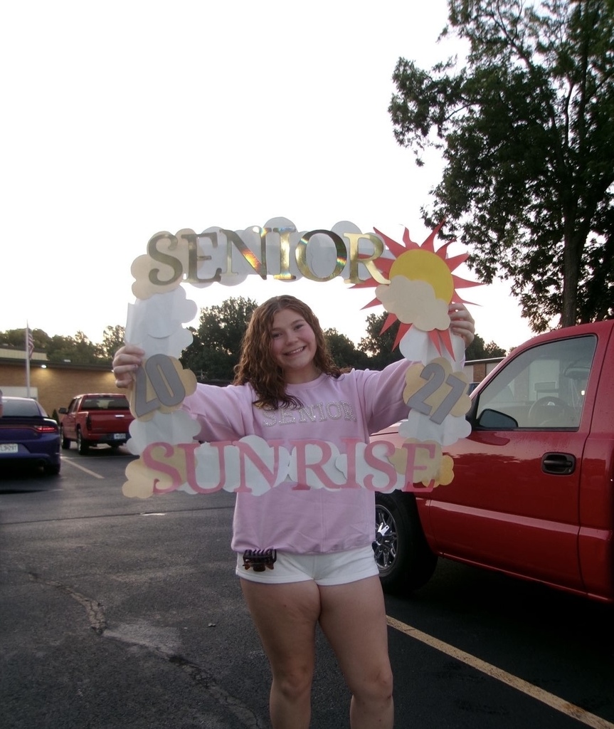 A smiling student in a pink “Senior” sweatshirt holds a colorful “Senior Sunrise 2027” frame in a parking lot.