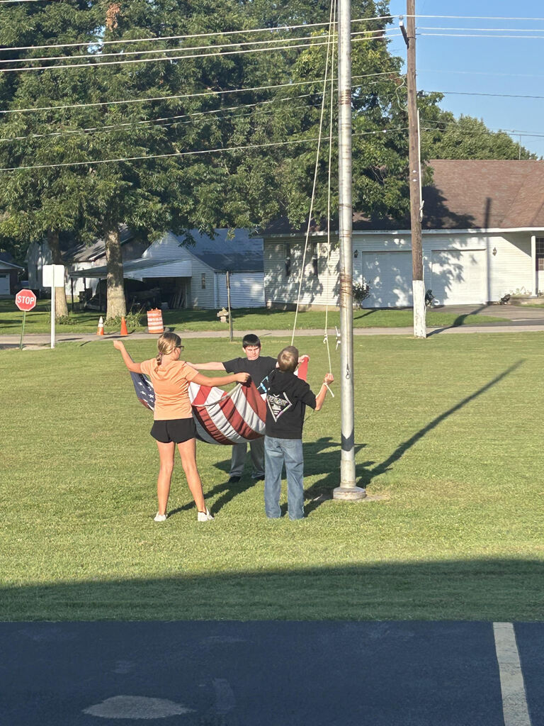 Three students work together to raise or lower an American flag beside a flagpole on a sunny school lawn.
