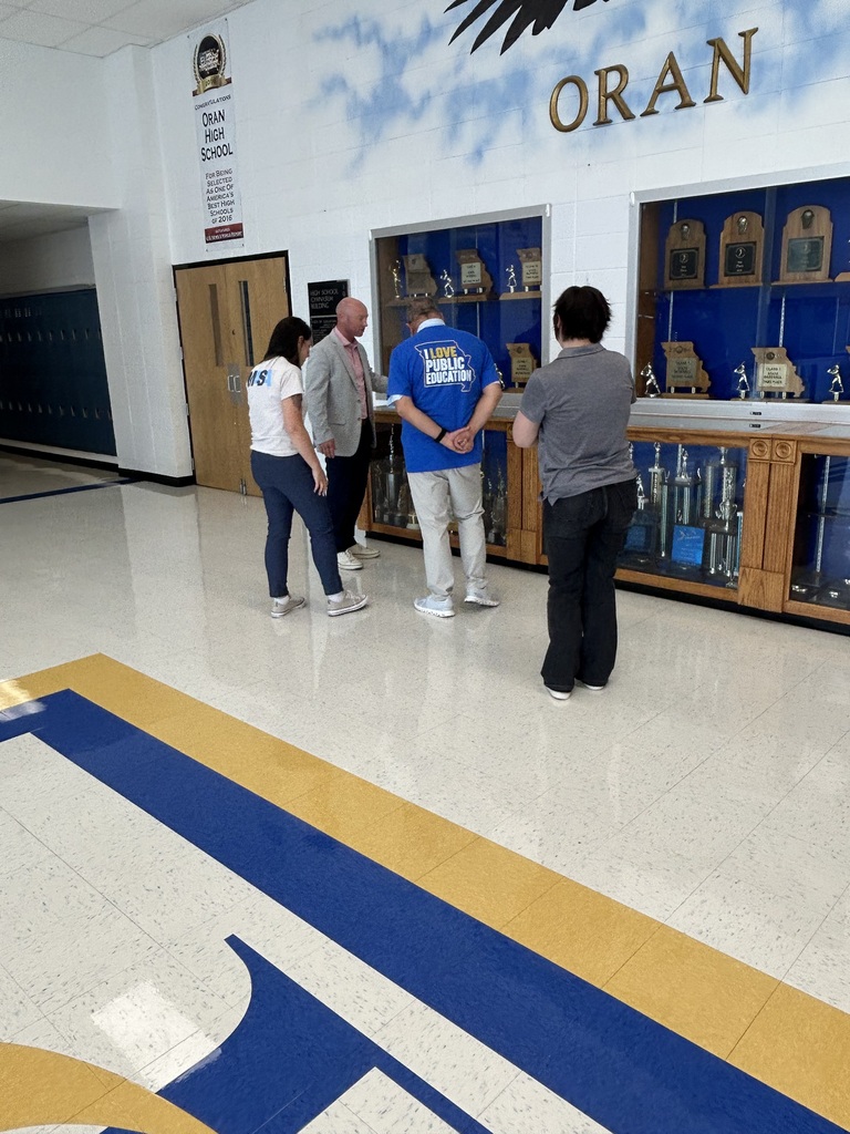 Four adults examine trophies and awards in display cases along an Oran High School hallway.
