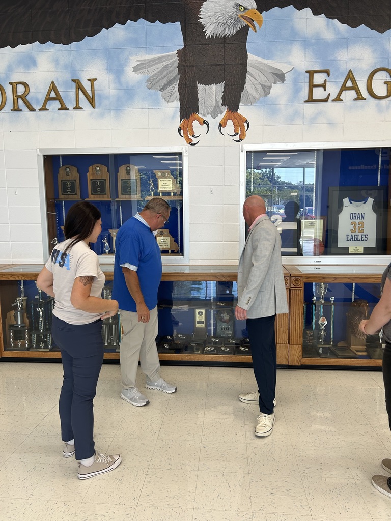 Three adults view trophies and memorabilia in a school display case beneath a large bald eagle mural.