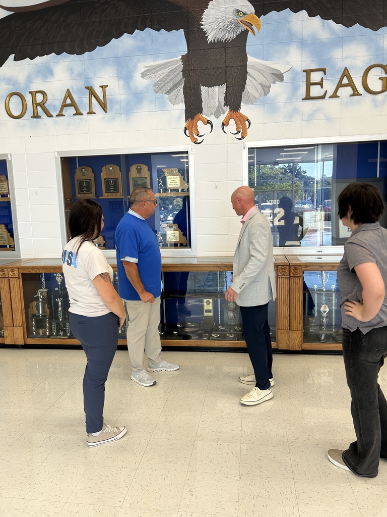 Four adults gather around trophy cases in a school hallway beneath a large mural of a soaring bald eagle.