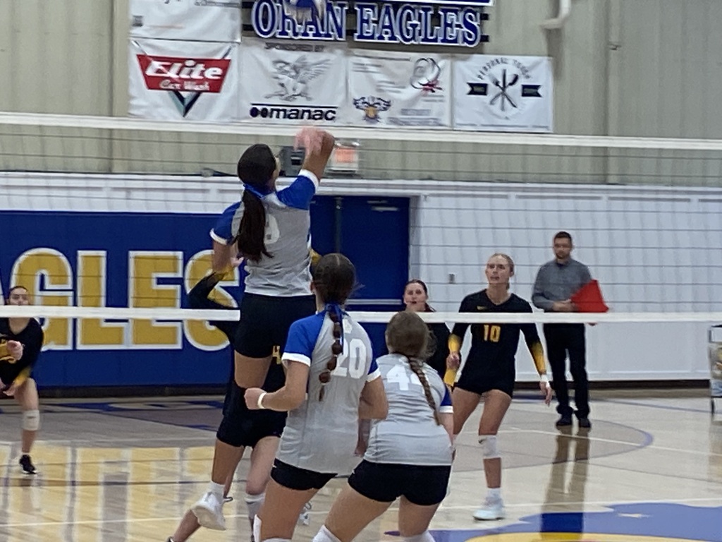Gray-and-blue volleyball player jumps near the net to play the ball as teammates and opponents prepare for the return.