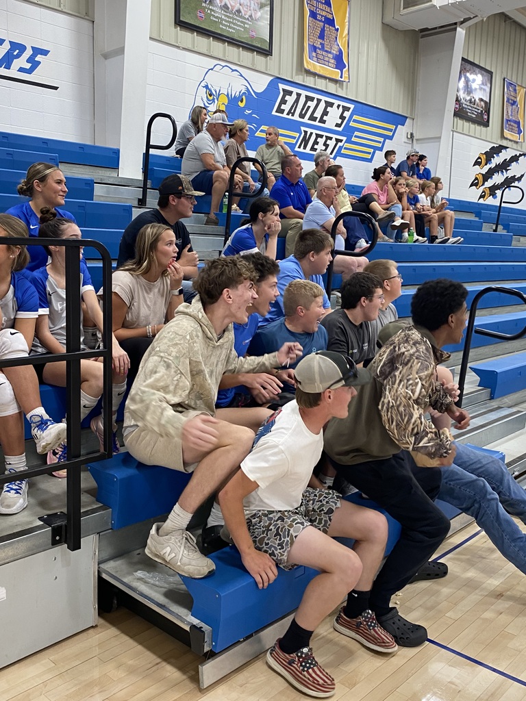 Excited spectators lean forward and cheer from bright blue bleachers during a volleyball match in the Eagles’ Nest gym.