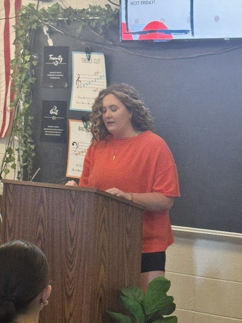 Young woman in a red shirt speaks from a wooden podium in a decorated classroom.