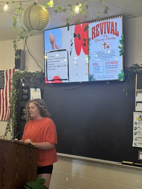 Young woman speaks at a podium beneath a screen displaying an FCA Morning Devotions revival slide.