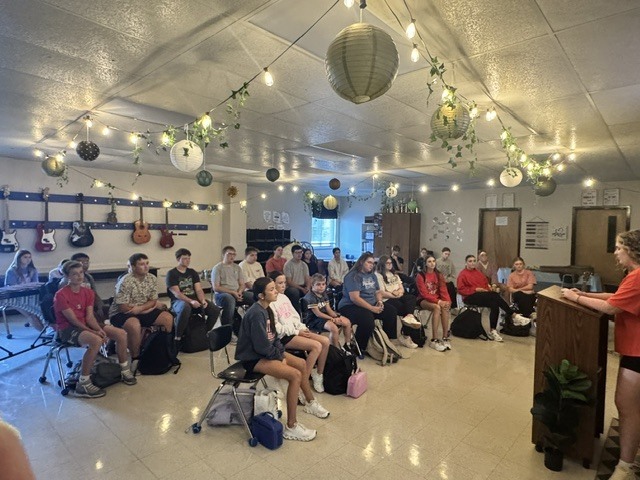 Students sit in rows listening to a speaker at a podium in a music classroom decorated with lights.