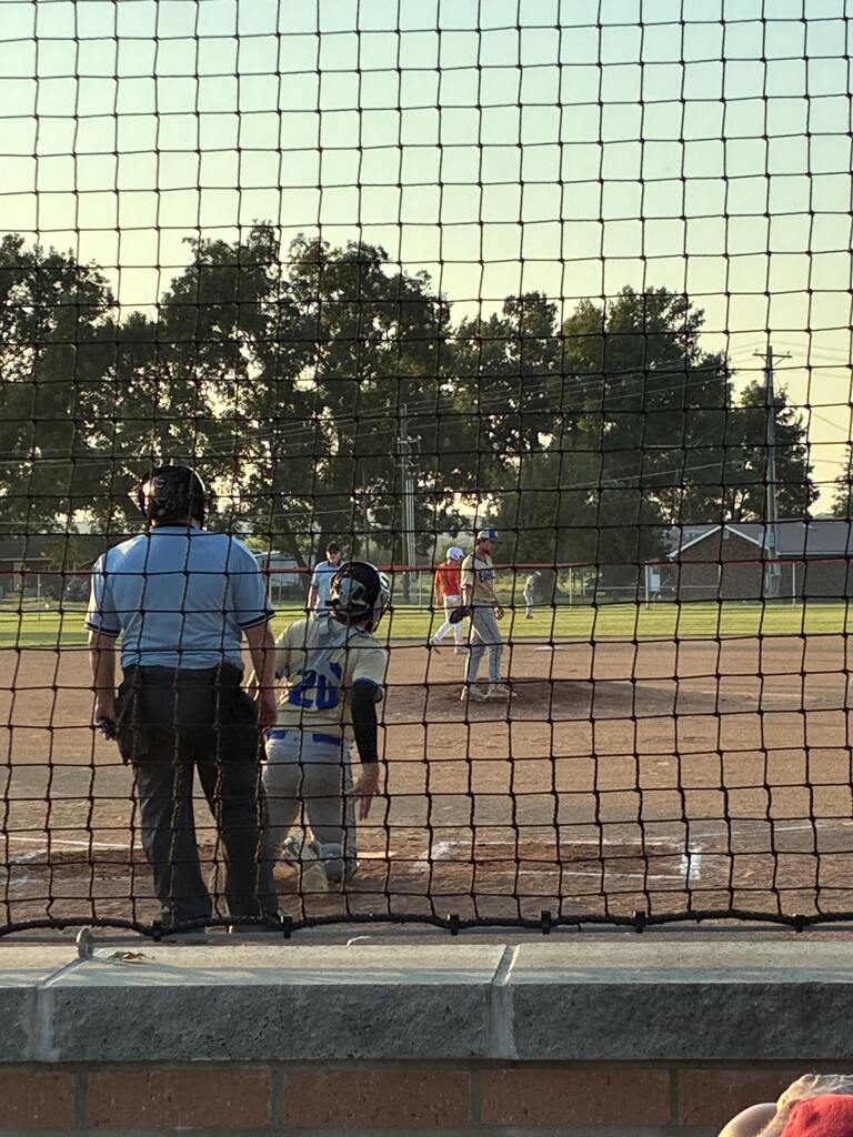 Catcher and umpire stand behind home plate as players prepare for the next pitch during a sunset baseball game.