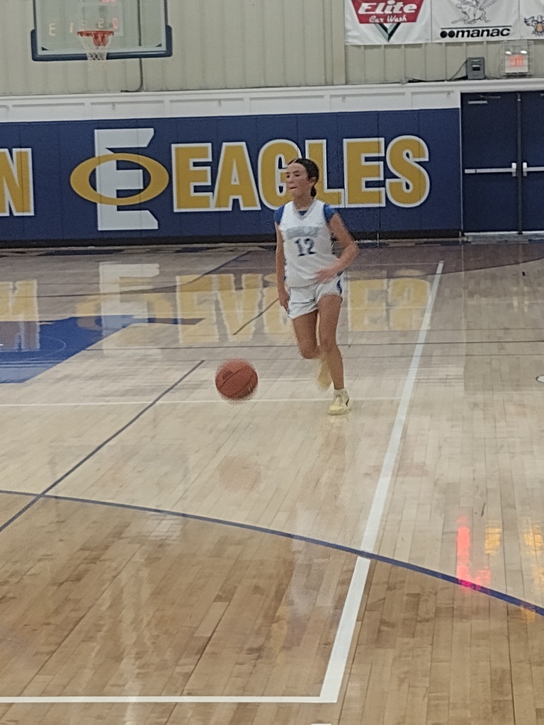 Image 3: A basketball player wearing white number 12 dribbles up the court with the Eagles logo behind her.