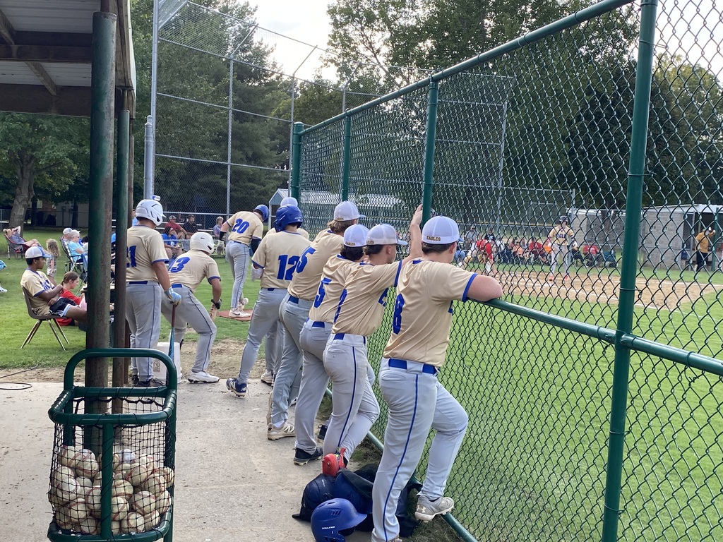 Baseball team in tan jerseys leaning up against a fence at a baseball field.