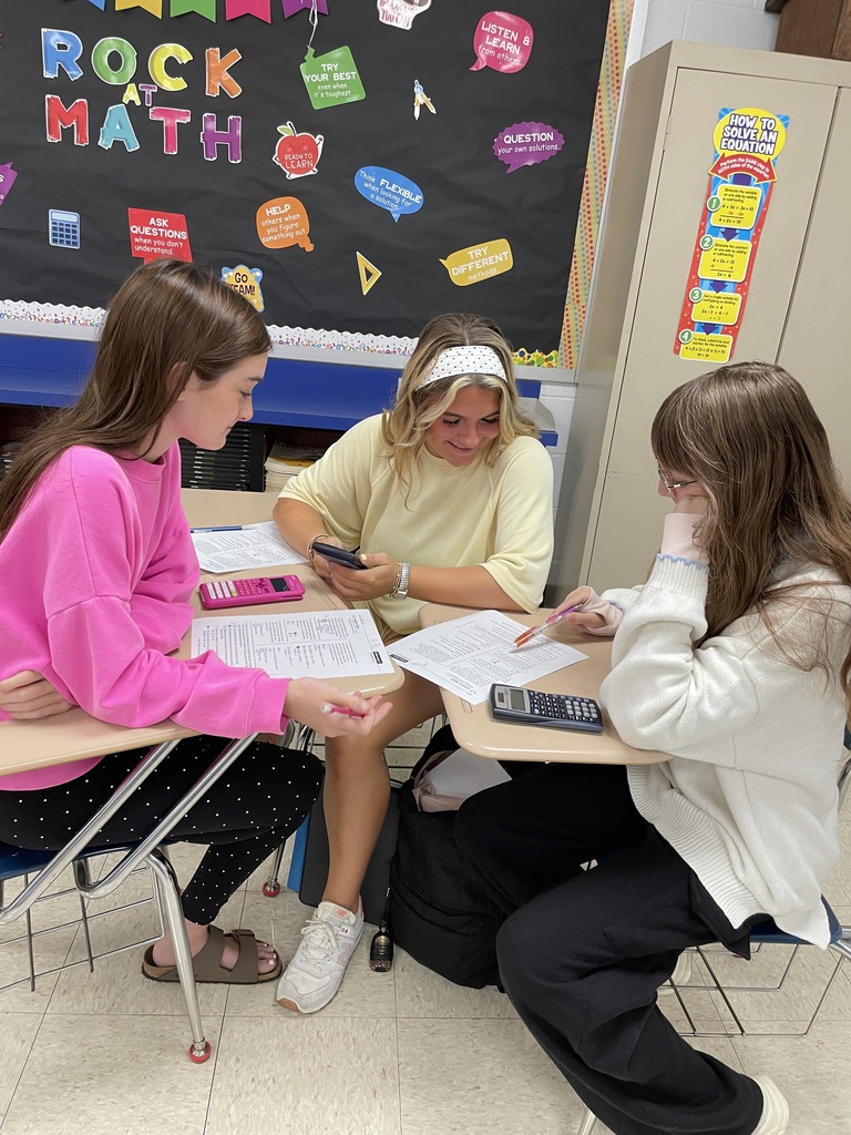 Three students work together on a math assignment at their desks, using worksheets and calculators. A colorful “Rock at Math” bulletin board is displayed behind them.