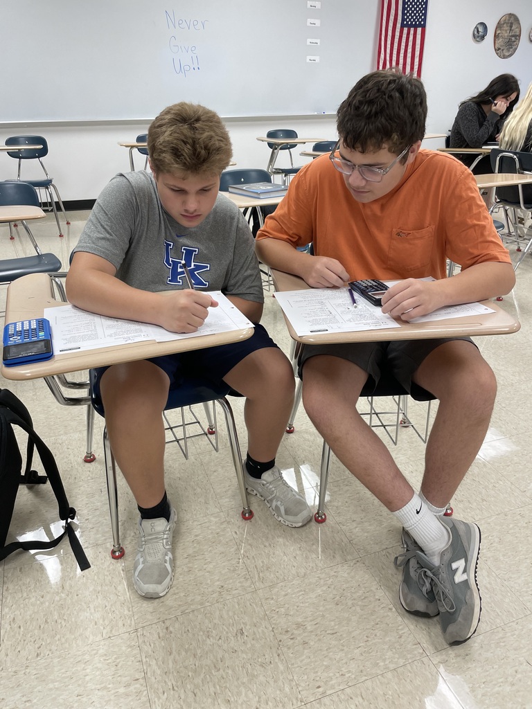 Two students sit side by side in a classroom, working together on math worksheets with calculators. A whiteboard behind them reads, “Never Give Up!!”