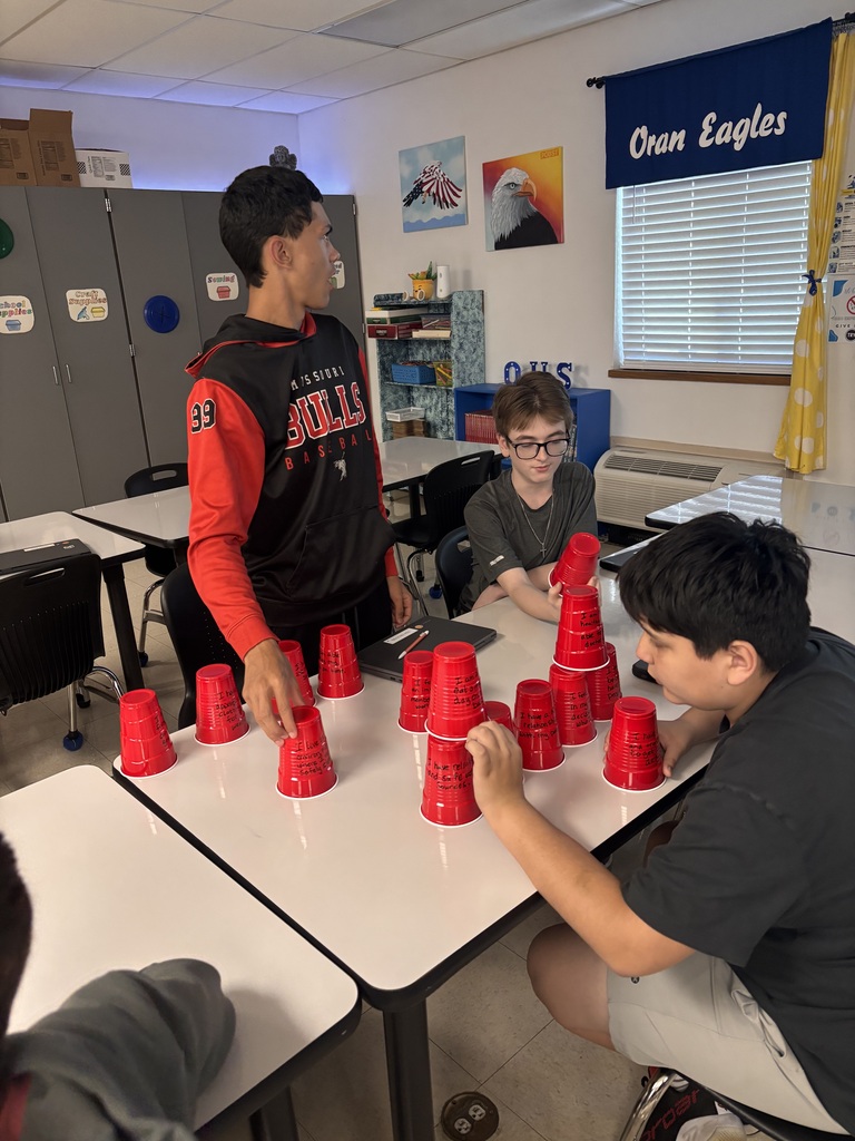 Three students collaborate at a classroom table, arranging red cups with handwritten messages into small stacks.