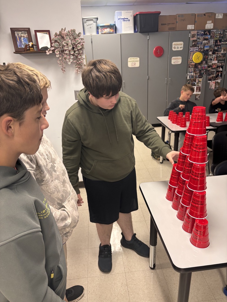 Three students examine a tall pyramid of red cups covered with handwritten messages during a classroom activity.