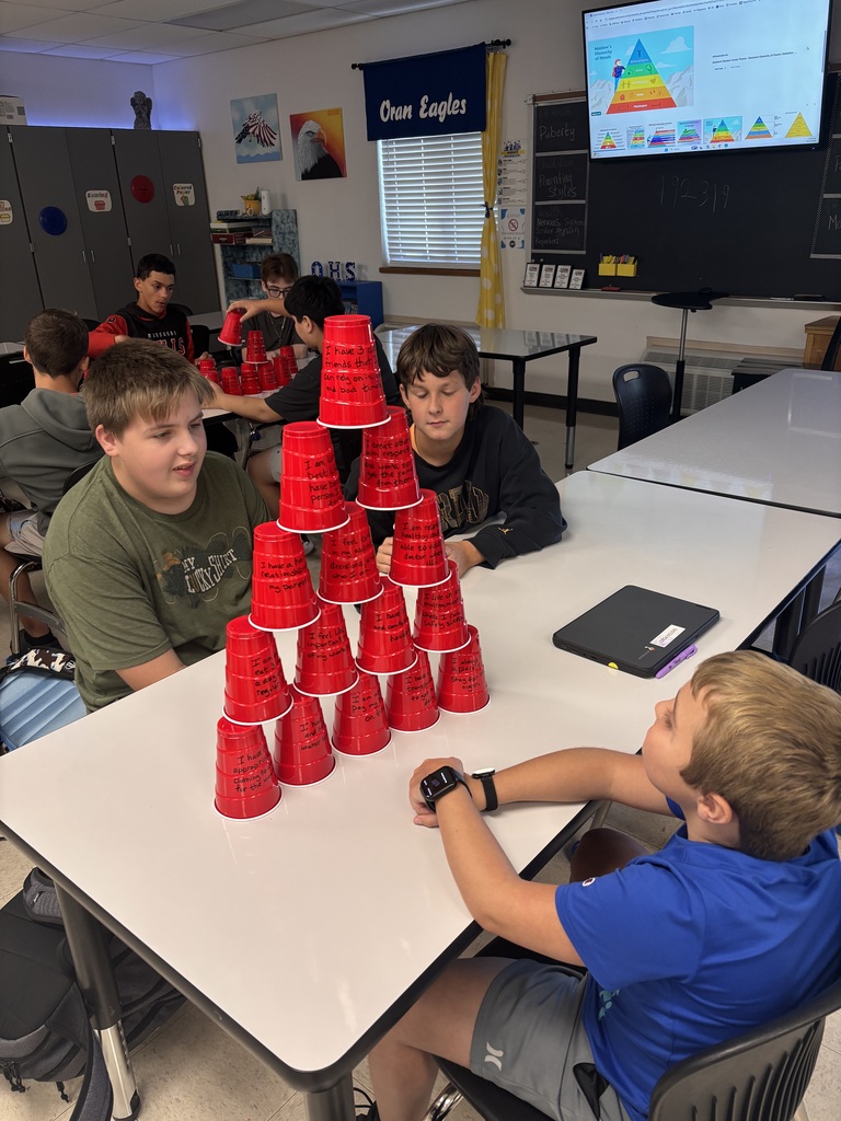 Students sit around a classroom table, working together to build a tall pyramid from red cups covered in handwritten messages.