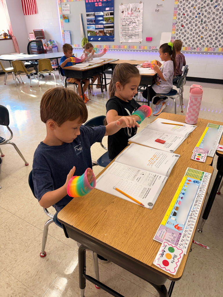 Elementary students sit at classroom desks, stretching colorful rainbow springs between their hands during a lesson.