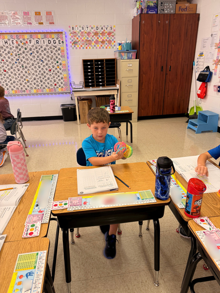 A student seated at his classroom desk holds a coiled rainbow spring above an open workbook.