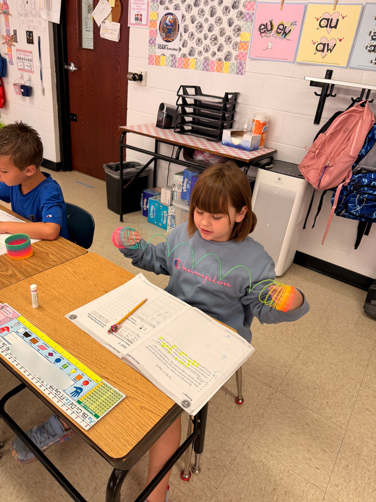 A student at her classroom desk stretches a rainbow spring between both hands above an open workbook.