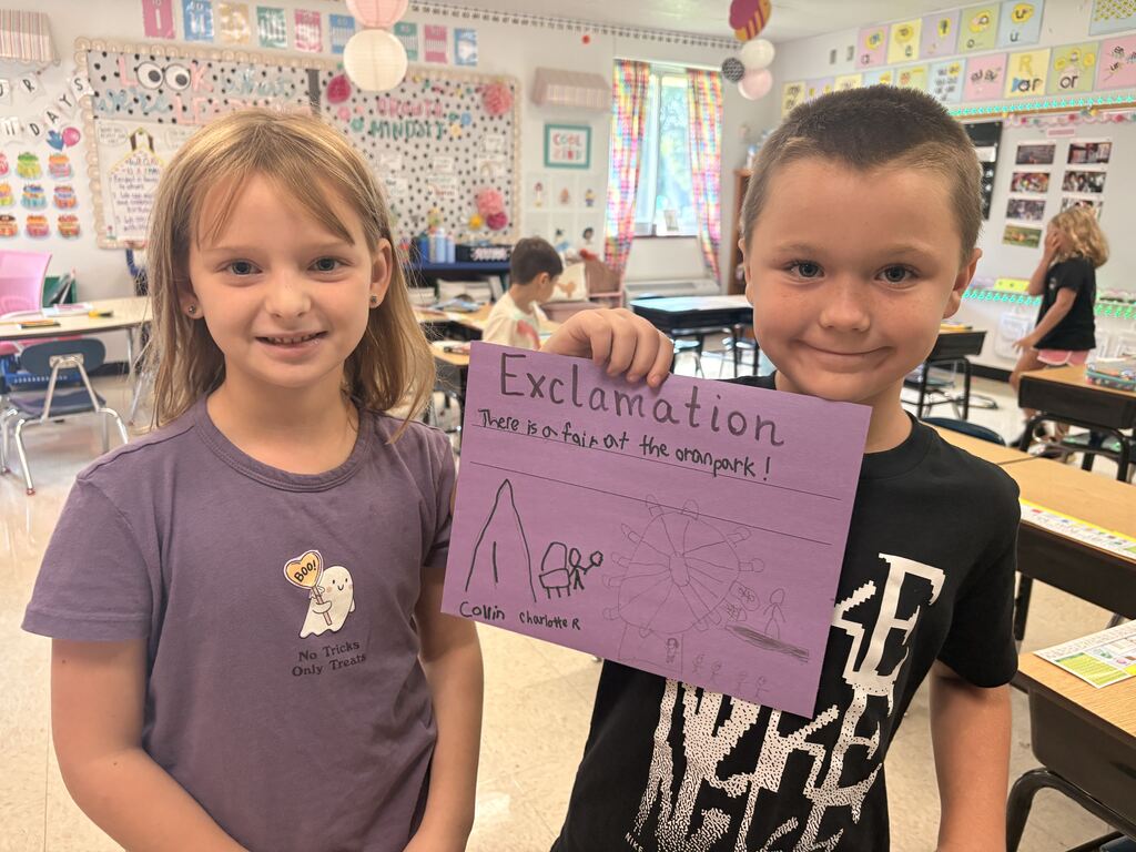 Two smiling students display a purple “Exclamation” poster about a fair at the orange park, illustrated with a Ferris wheel.