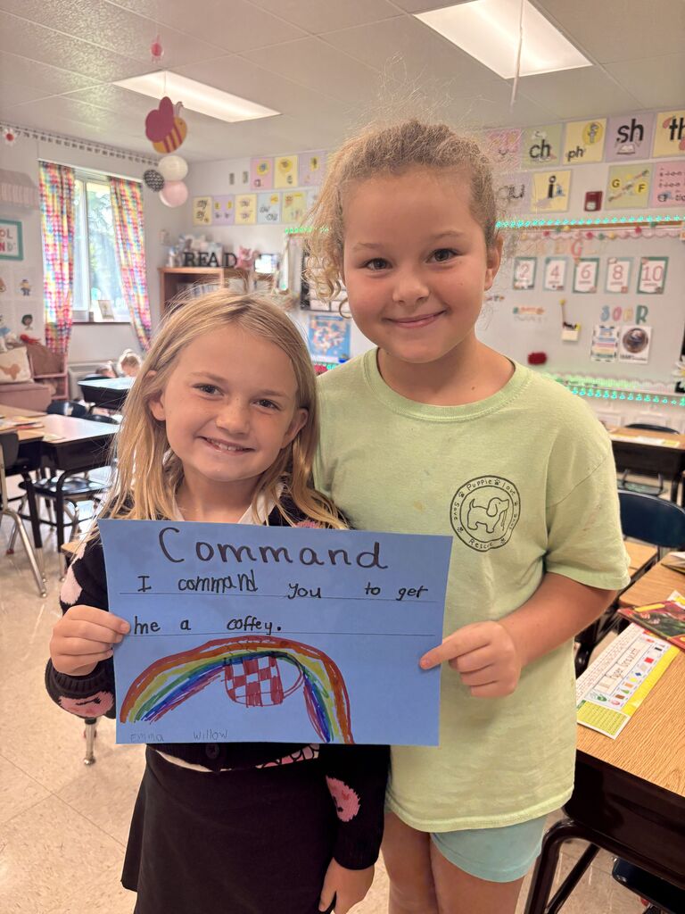 Two smiling students hold a blue “Command” poster with a rainbow drawing in a colorful classroom.