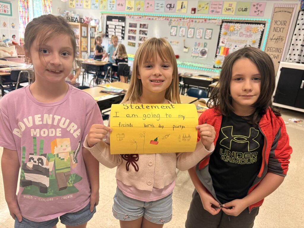 Three students pose together as the center student holds a yellow “Statement” poster about a friend’s birthday party.