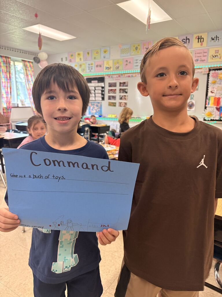 Two students stand together while one holds a blue “Command” poster reading, “Give me a bunch of toys,” with small drawings.