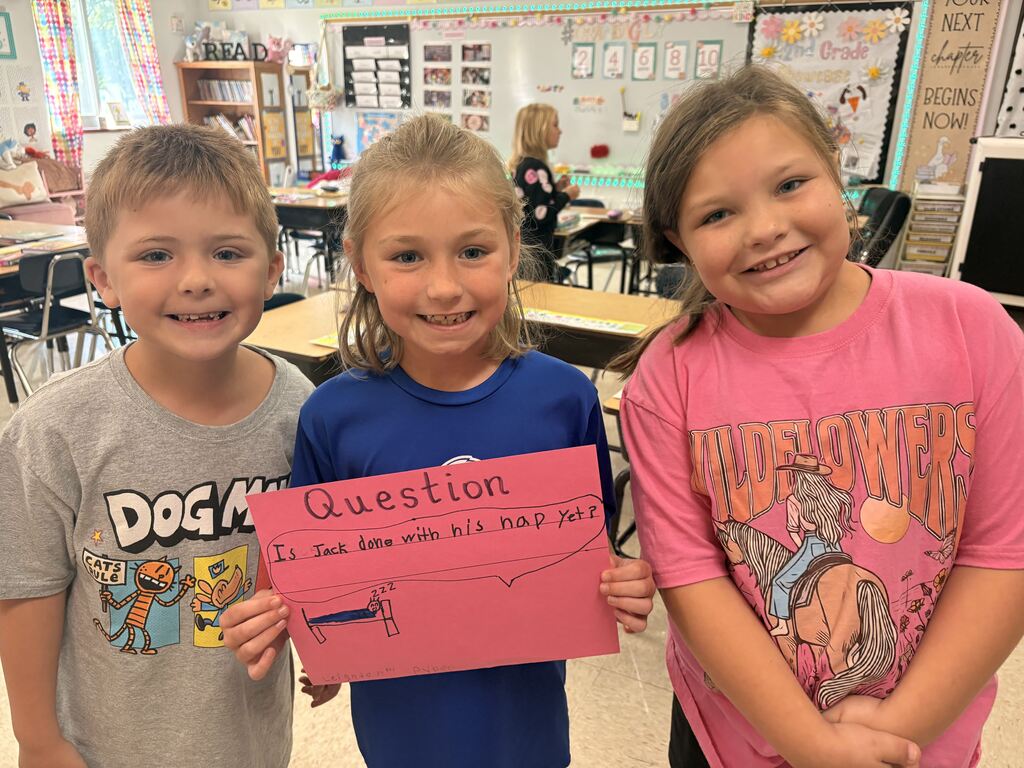 Three smiling students display a pink “Question” poster reading, “Is Jack done with his nap yet?” with a sleeping figure.