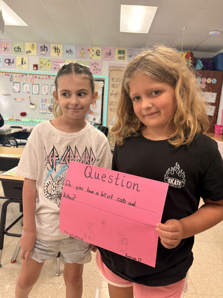 Two students pose with a pink “Question” poster asking, “Do you have a lot of cats and kittens?”