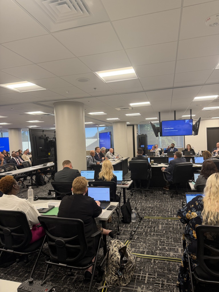 Education officials and attendees gathered around tables with laptops during a formal meeting and presentation.