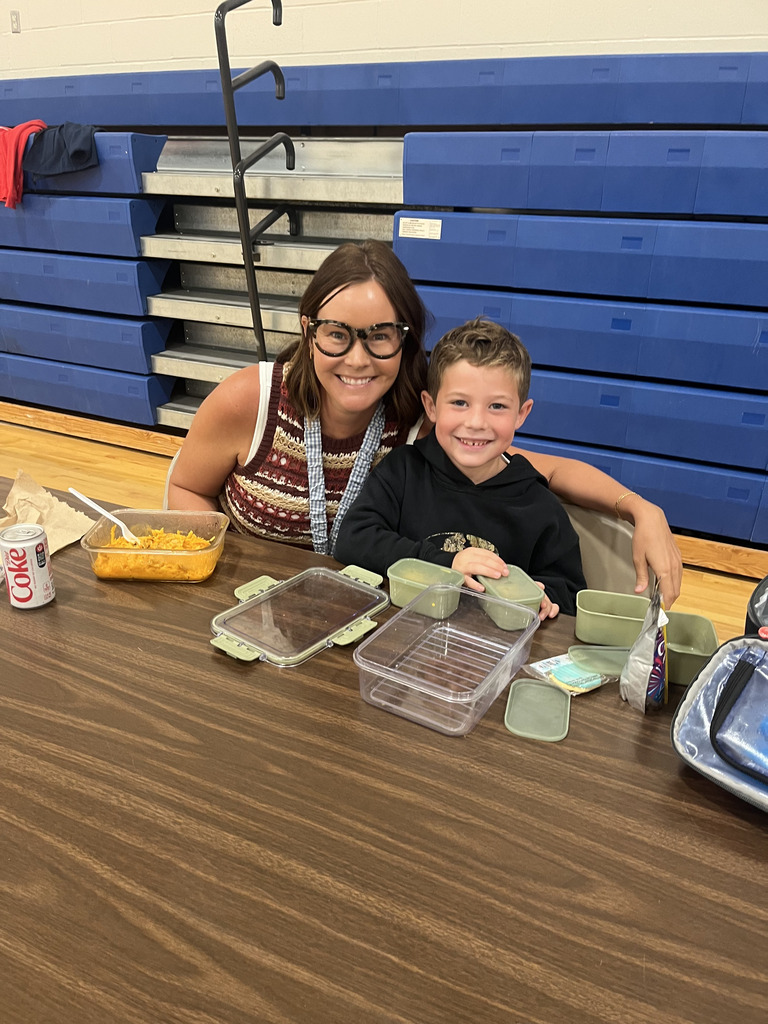 Student sitting next to teacher smiling with lunch food on a table in school cafeteria.