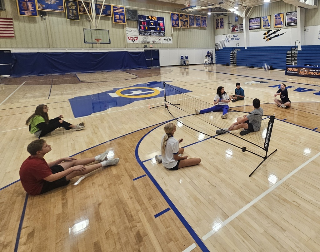 Students sit on both sides of a low net during a seated volleyball game, with one player preparing to hit the ball.