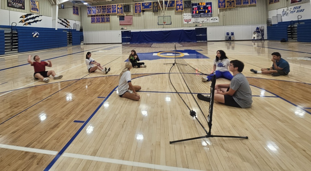 Students sit on both sides of a low net during a seated volleyball game, with one player preparing to hit the ball.