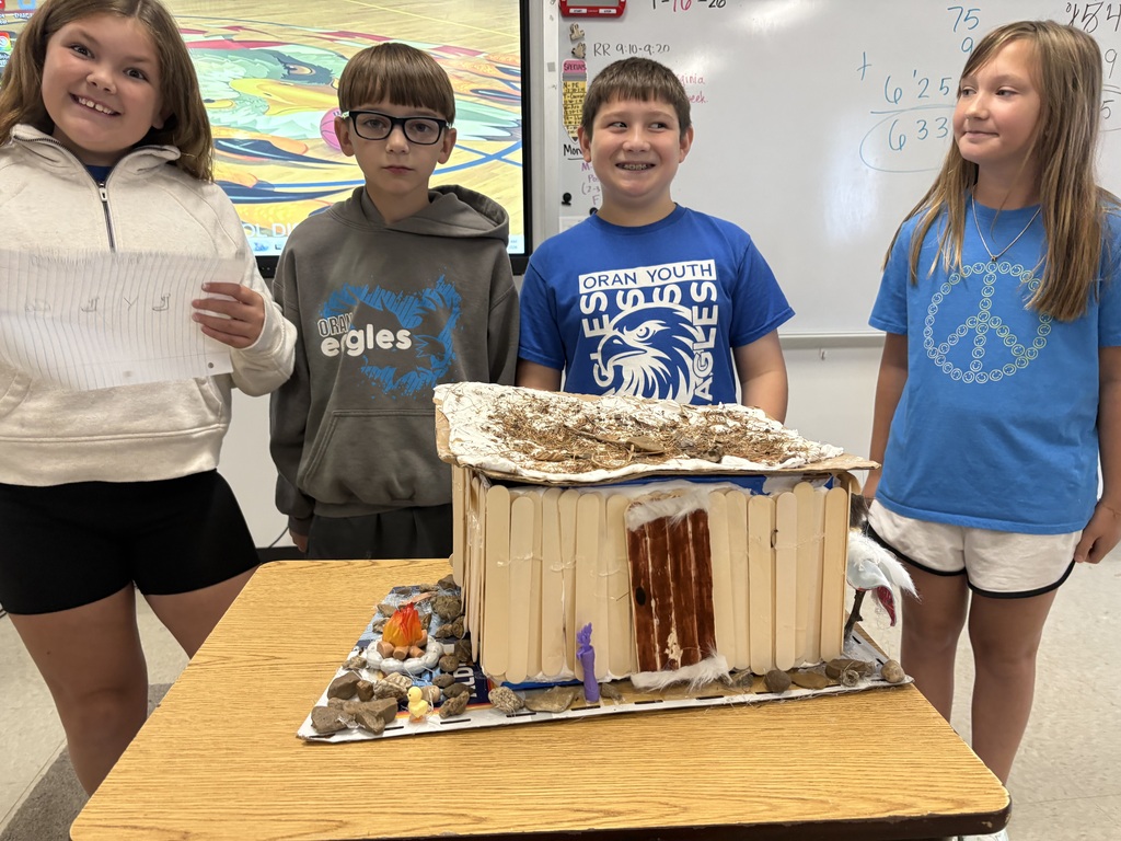 Four students stand behind a handmade model shelter built with craft sticks, rocks, and natural materials.