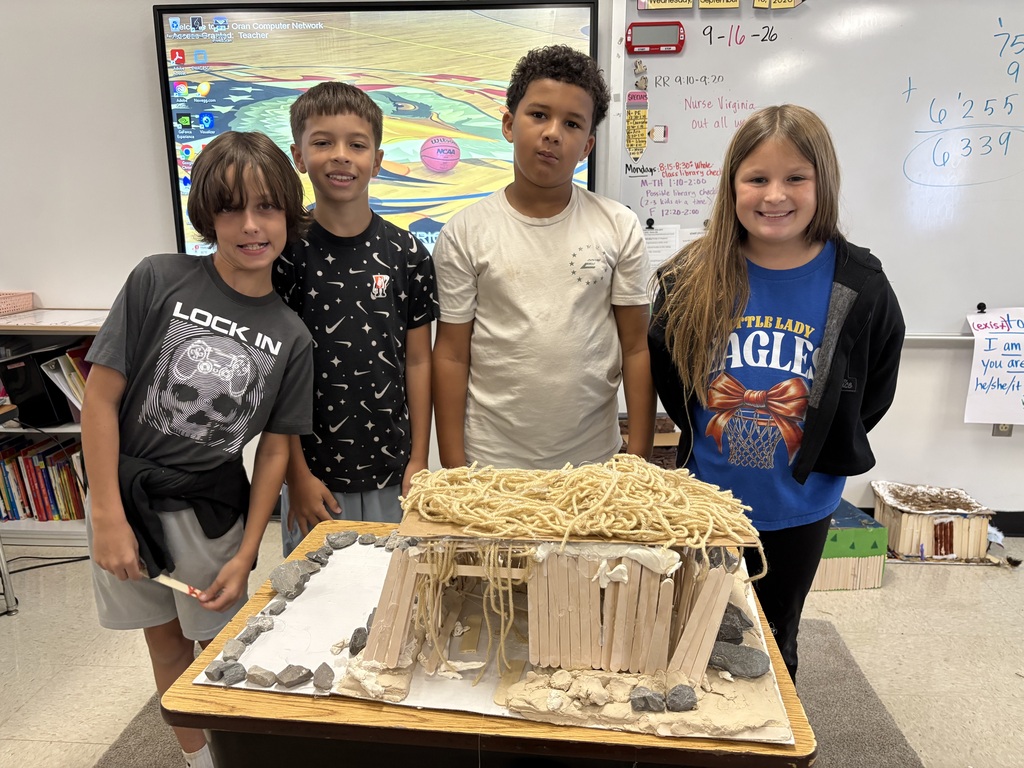 Four students stand behind a model shelter made from craft sticks and yarn, surrounded by rocks and clay.