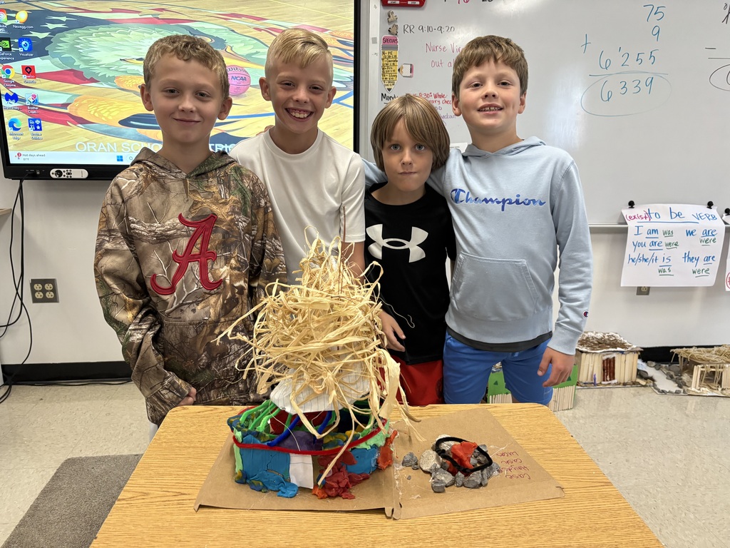 Four students pose behind a colorful model shelter topped with straw, with a small stone-ringed fire beside it.