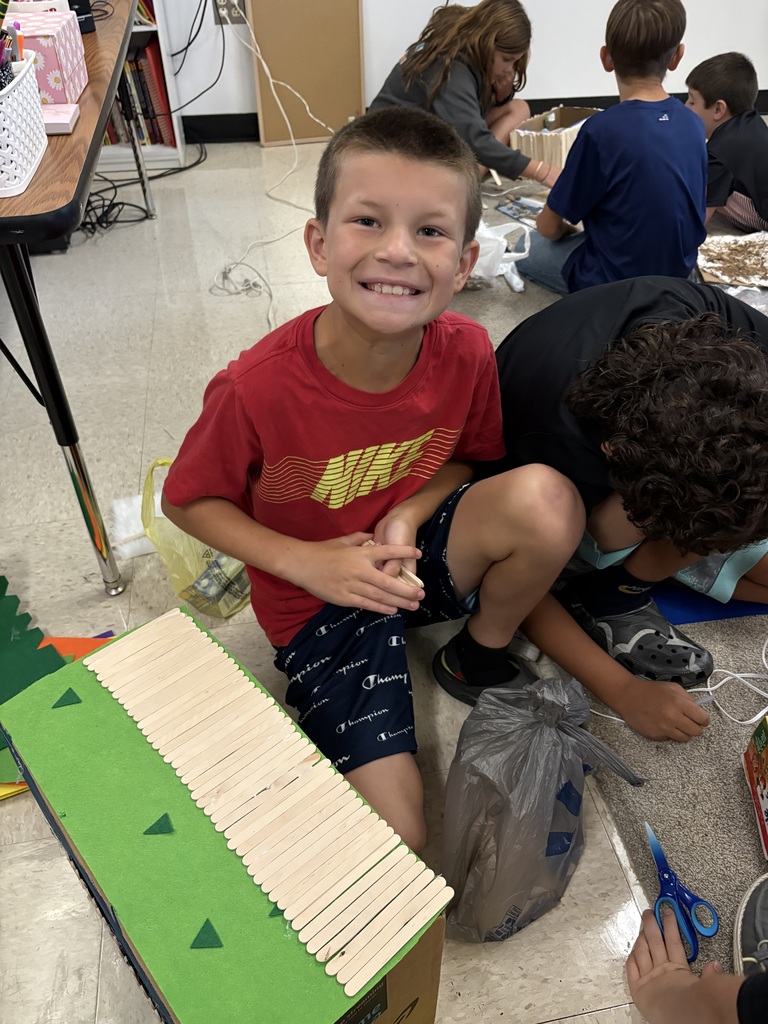 Smiling student works with classmates to build a model structure from craft sticks and other classroom materials.