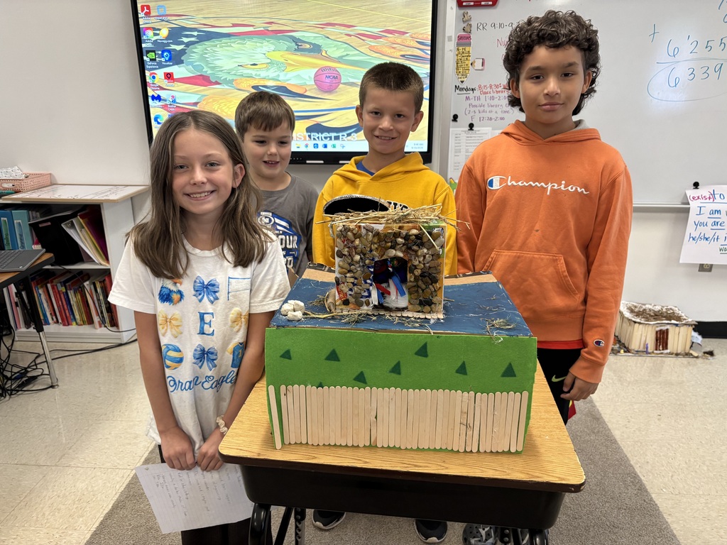 Four students pose behind a colorful model shelter decorated with stones, straw, and craft sticks.