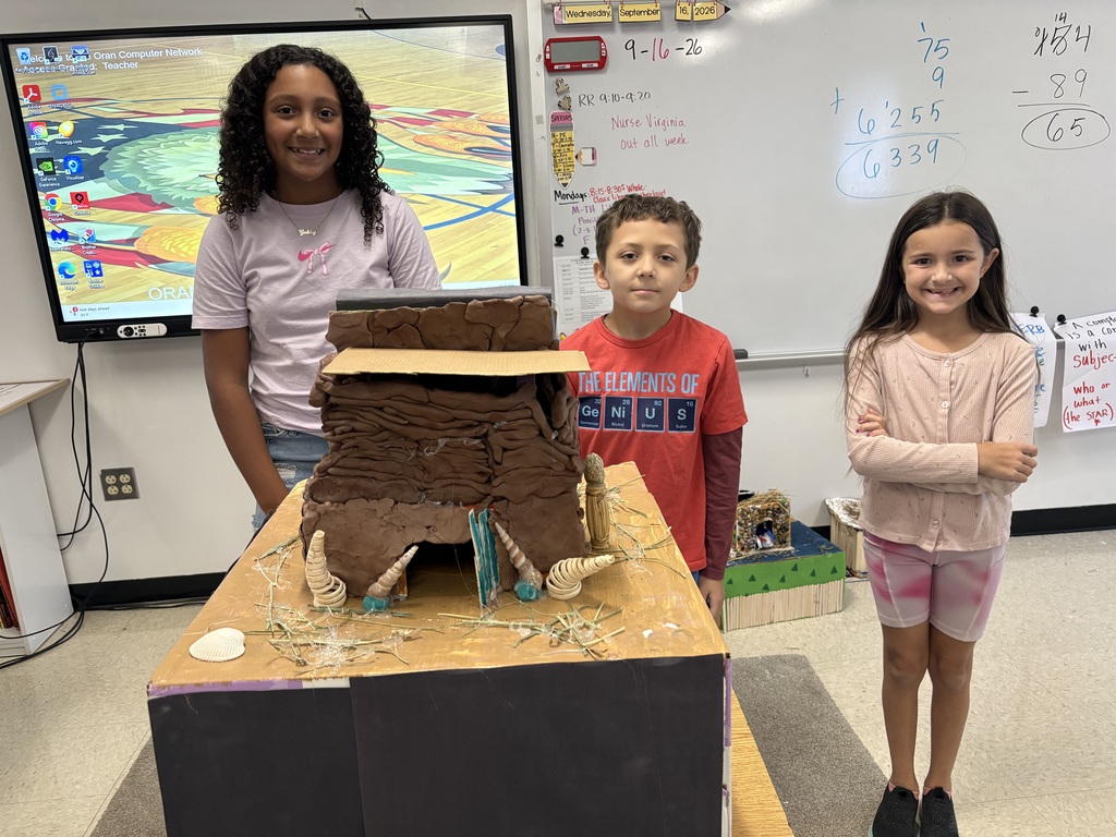 Three students stand behind a large handmade model shelter constructed from clay, cardboard, and natural materials.
