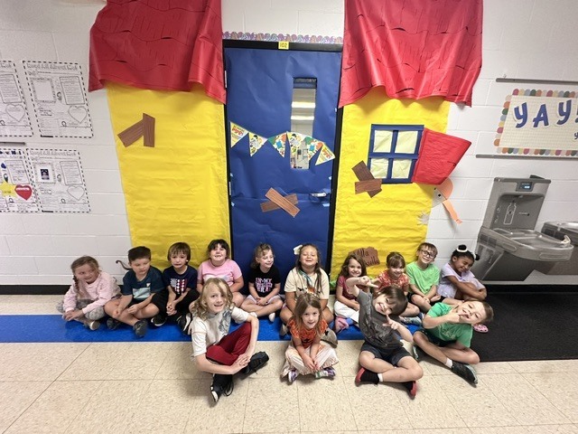 A group of smiling young students sit together in a school hallway beneath a colorful, house-themed classroom doorway.