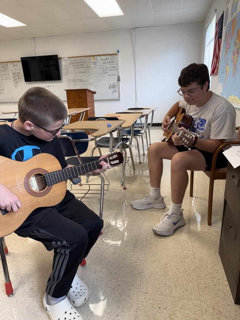 Two students sitting in a school classroom playing acoustic guitars.