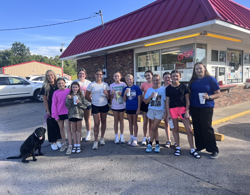 Group of young girls and a dog standing outside of a ice cream shop smiling and holding cups.