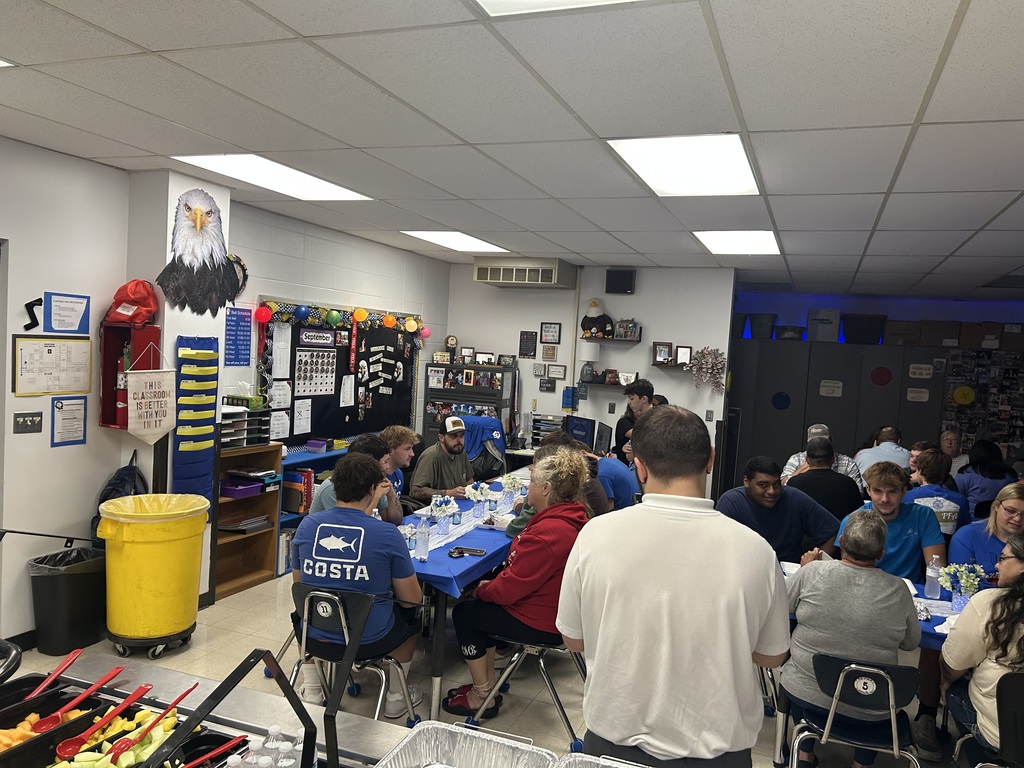 Students, families, and staff share breakfast at crowded classroom tables, with fruit and drinks served nearby.