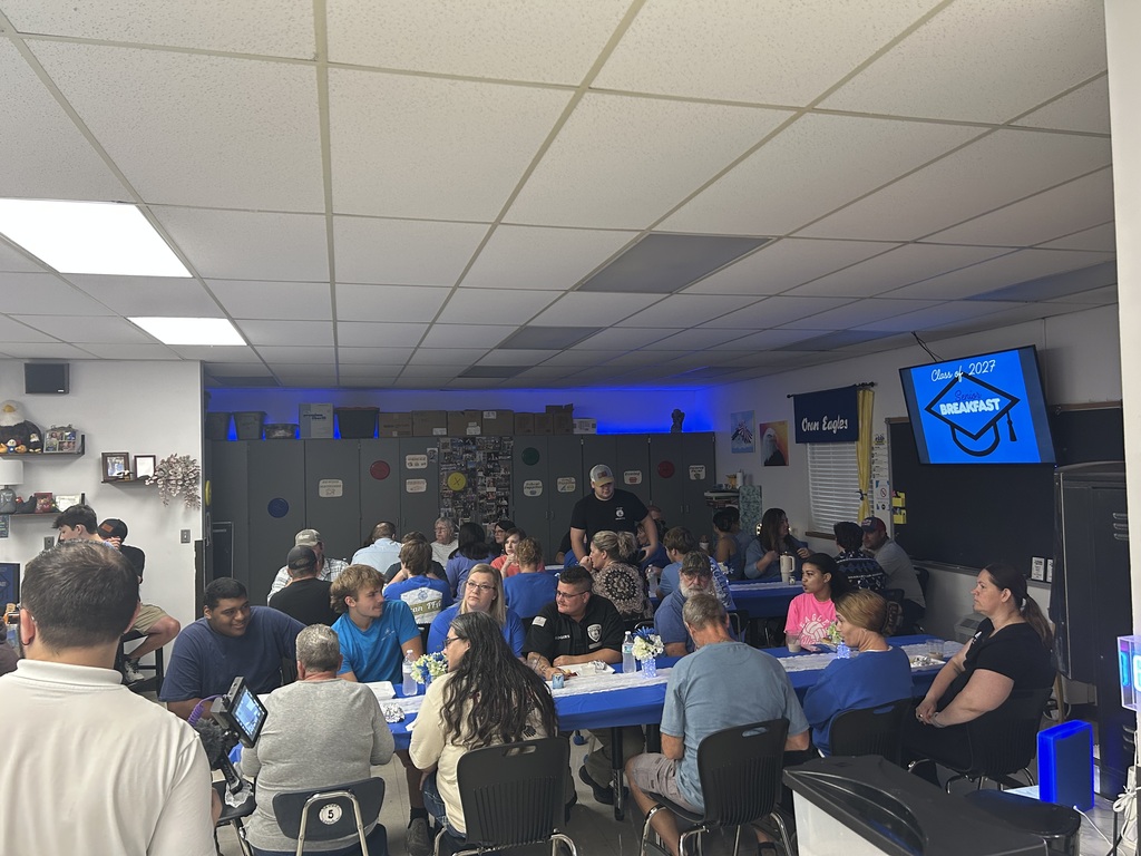 Students and adults gather around blue-covered tables in a classroom for a Class of 2027 senior breakfast.