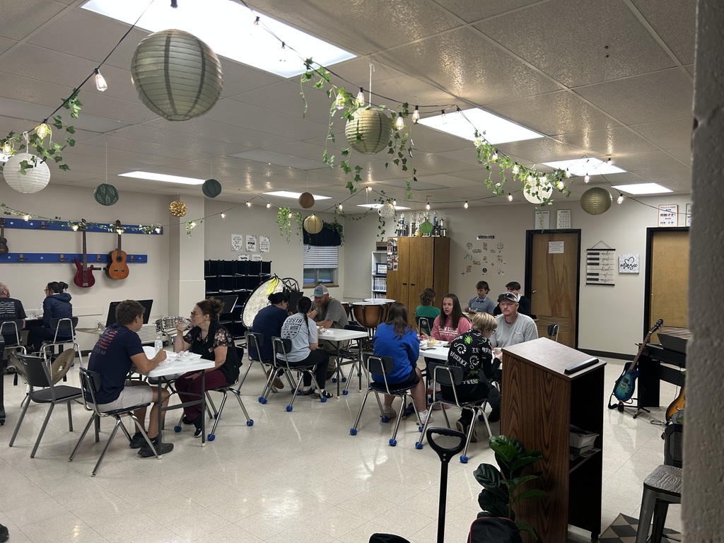Students and adults eat and socialize at tables in a music classroom decorated with string lights and hanging greenery.