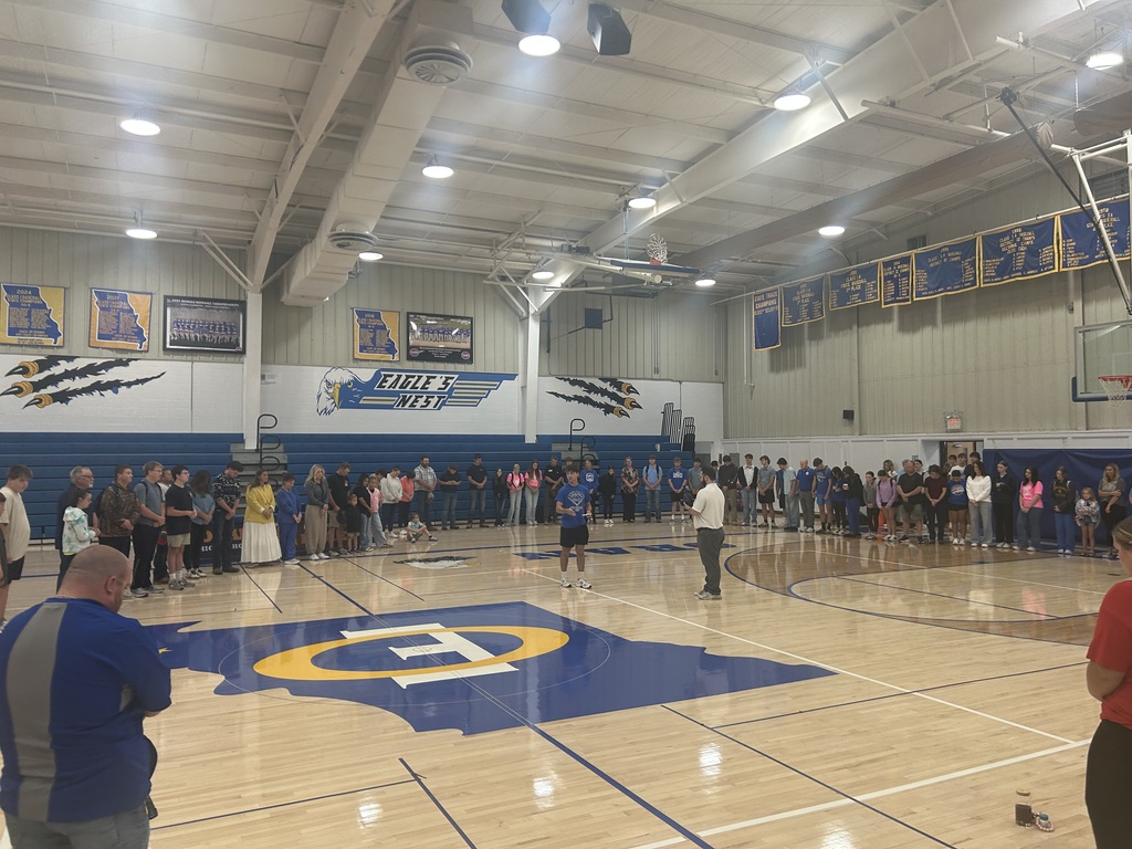 Students, families, and staff stand in a large circle on a high school gym floor beneath blue-and-gold banners.