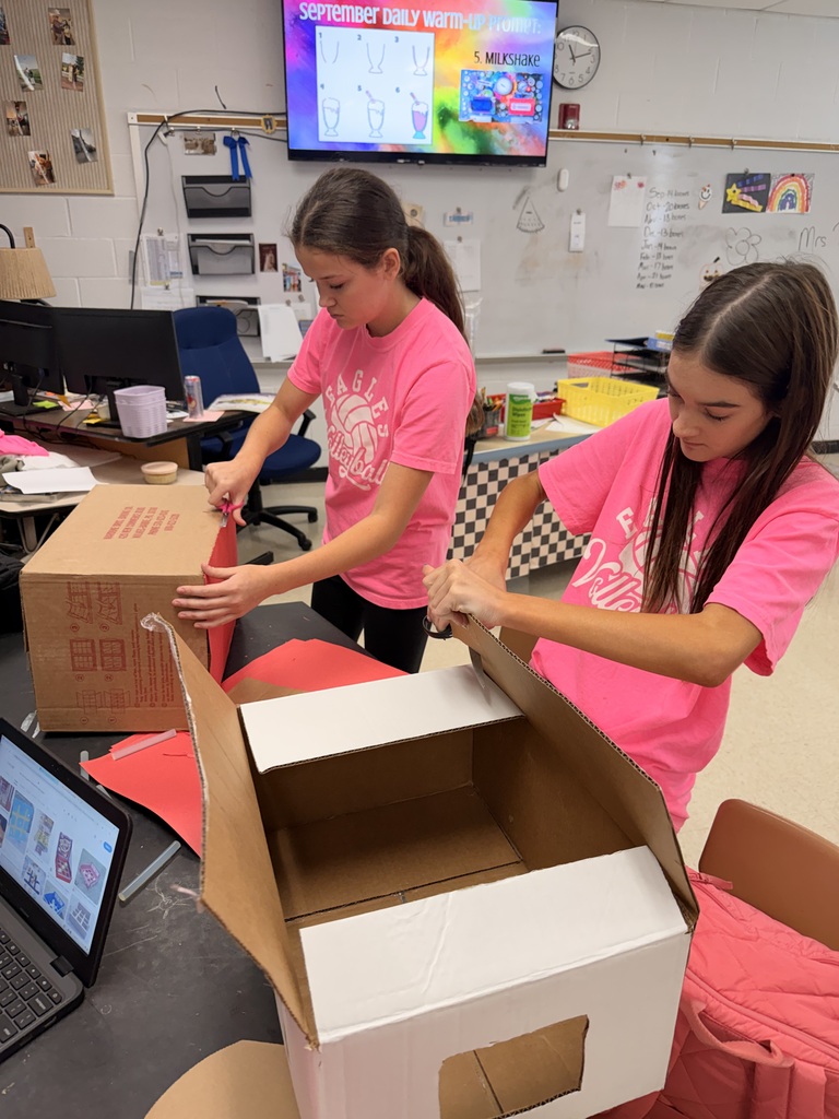 Two students in bright pink shirts cut and shape cardboard boxes together during a classroom project.