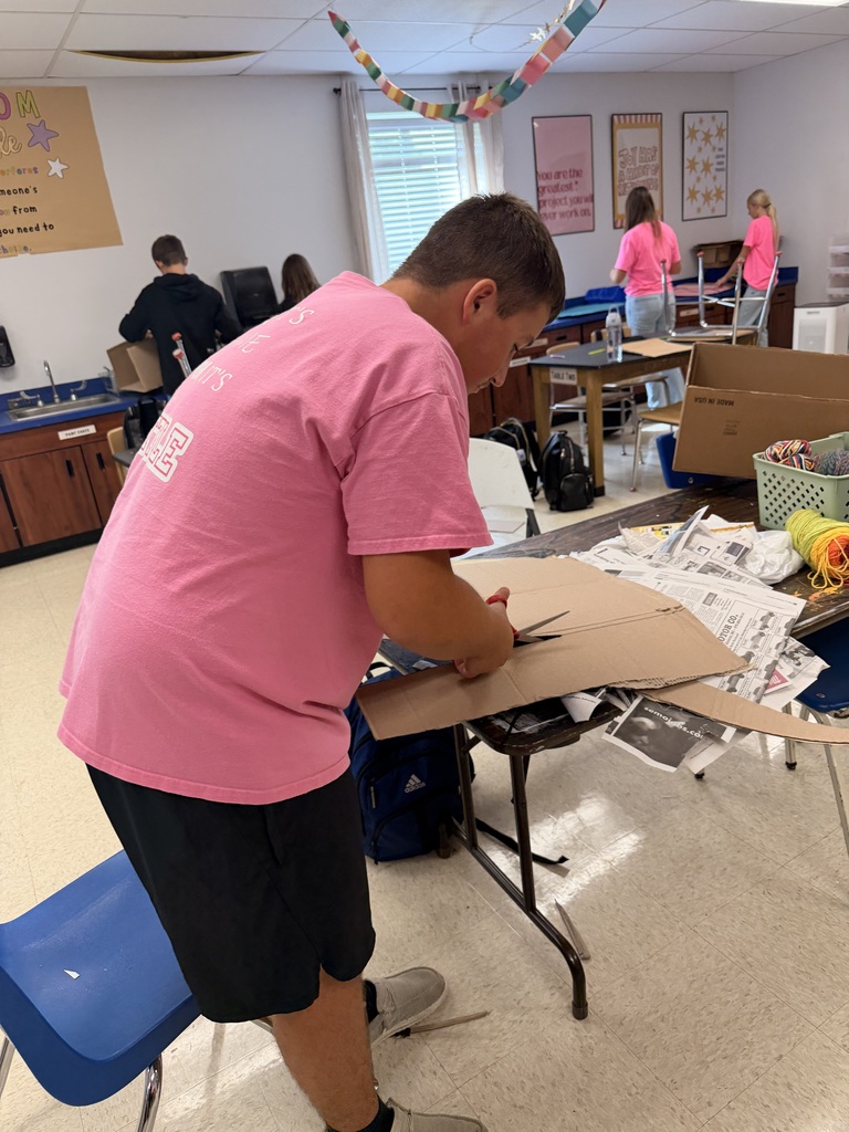 Student in a pink shirt cuts cardboard at a classroom table while classmates work on projects in the background.