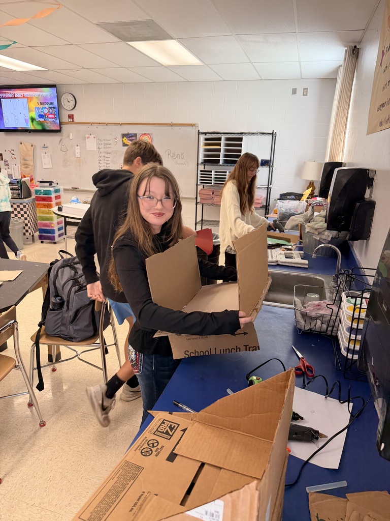 Student holds a partially assembled cardboard structure while classmates work on projects around the classroom.